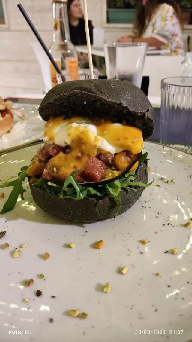 The image prominently features a black bun burger, likely tuna, served on a white plate. The focus is sharp on the burger, which appears photo-realistic with accurate colors. The background is slightly blurred but includes a glass and other elements, and there are some crumbs on the plate.