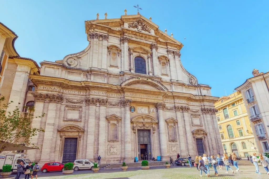 A grand, ornate Baroque church facade under a clear blue sky, with some people and cars in the foreground.