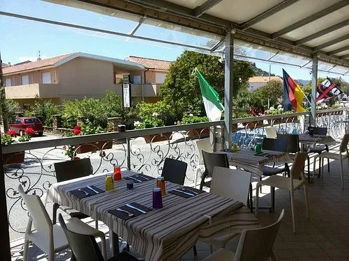 An outdoor dining patio of a restaurant or cafe, featuring multiple tables set with striped tablecloths, place settings, and colorful bottles. A decorative metal railing is visible in the foreground, separating the patio from a street. In the background, national flags (Italian and German) are flying, with buildings, green trees, and a clear blue sky.