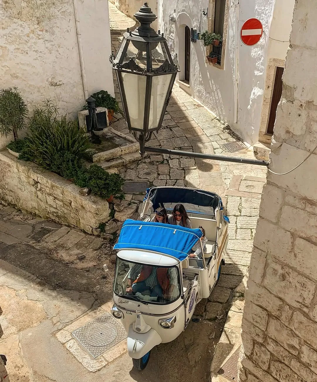 An elevated view of a white three-wheeled vehicle with two passengers driving down a narrow, sunlit cobblestone street lined with white buildings in an old European town, featuring a prominent black lamppost.