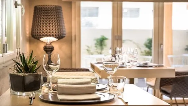 The image displays a well-composed and aesthetically pleasing dining table setting within what appears to be a restaurant or upscale interior. The foreground elements, including glasses, plates, and napkins, are sharp and in focus. Natural light enters from a large window in the background, providing good illumination. The colors are accurate and contribute to the photo-realistic quality of the image. The composition effectively draws attention to the table setting while providing a pleasant, blurred background.