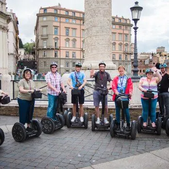 An outdoor shot of a group of tourists riding Segways in a city, with historic buildings and a large column in the background.
