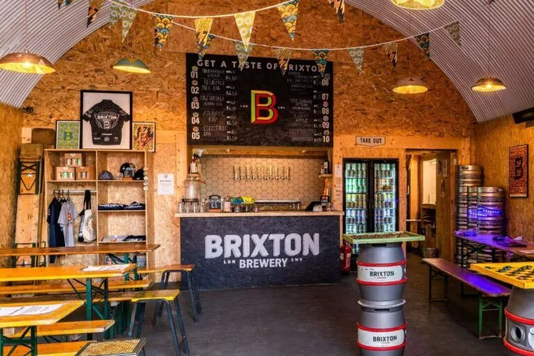 The image provides a clear view of a brewery taproom, featuring a well-composed shot of the bar area, menu board, and seating. The lighting is warm and generally even, contributing to an inviting atmosphere. The image is photo-realistic and the main elements, such as the bar and menu, are in focus. However, some areas could benefit from improved sharpness, especially towards the foreground and edges, and there is a minor glare from the fridge on the right.