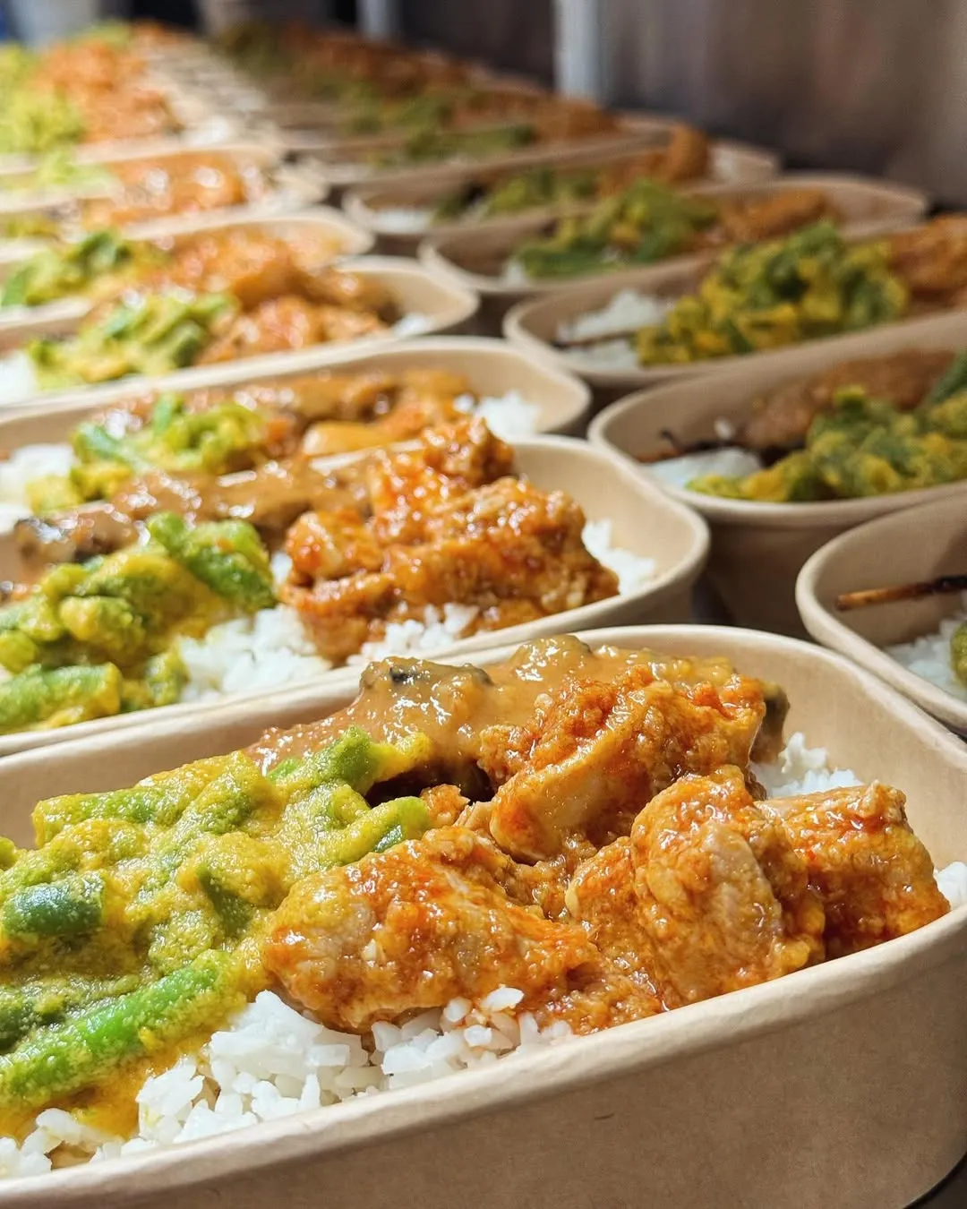 A close-up photograph showcasing multiple pre-packaged meal containers, each containing white rice, a vibrant green vegetable dish, and a sauced meat entree. The foreground meals are sharply in focus, creating a visually appealing depth of field that highlights the prepared food items.