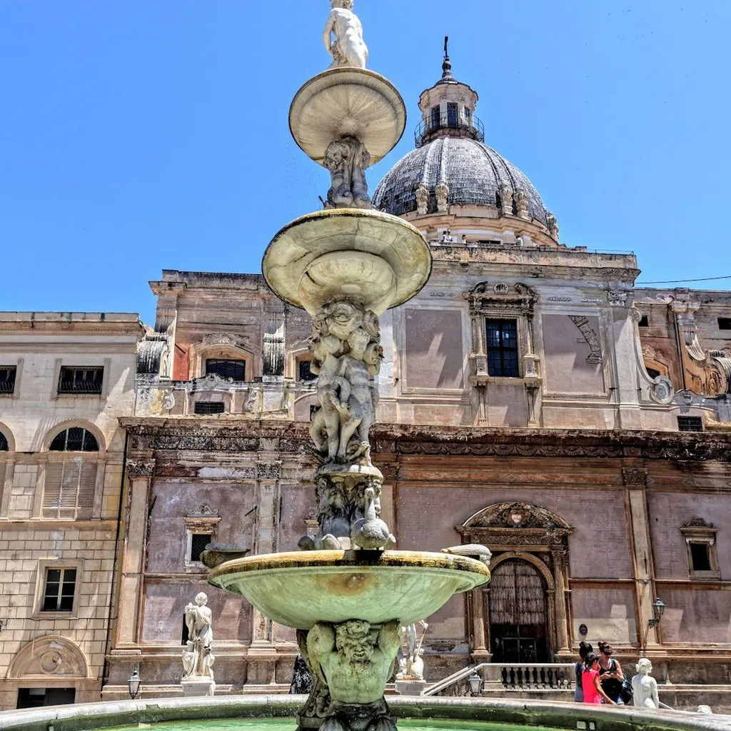 The image features a large, multi-tiered stone fountain as its central subject, set against the backdrop of an old, grand architectural building with a prominent dome under a clear blue sky. The scene is bathed in bright sunlight.