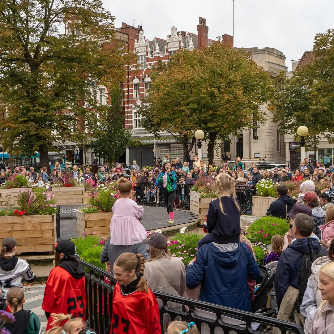 The image captures a vibrant outdoor event with a performer on a stage surrounded by an engaged audience, including children perched on shoulders. The scene is well-composed, featuring a mix of people, urban architecture, and trees under natural, overcast lighting. The colors are accurate and the image is highly photo-realistic, effectively conveying the lively atmosphere. Key elements, particularly the performer, are in good focus, contributing to the overall clarity.