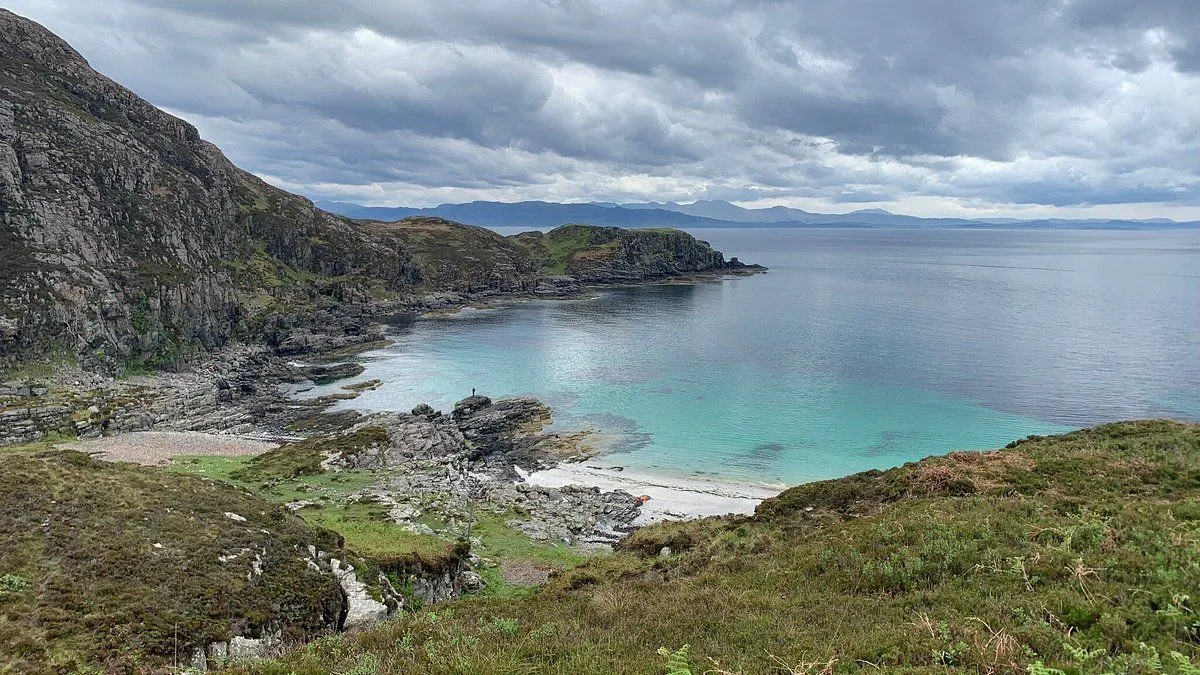 An editorial quality image depicting a scenic coastal bay on an overcast day. The composition is strong, balancing the sky, rugged coastline, and clear turquoise water. The lighting is even and natural, and the colors are accurate. While generally sharp, some areas like the foreground and distant elements could be crisper, and the overcast lighting results in a somewhat flat dynamic range.