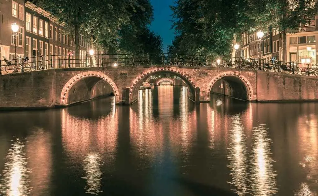 A picturesque night view of a canal with illuminated arched bridges, reflecting warm lights on the dark water, flanked by buildings and trees.