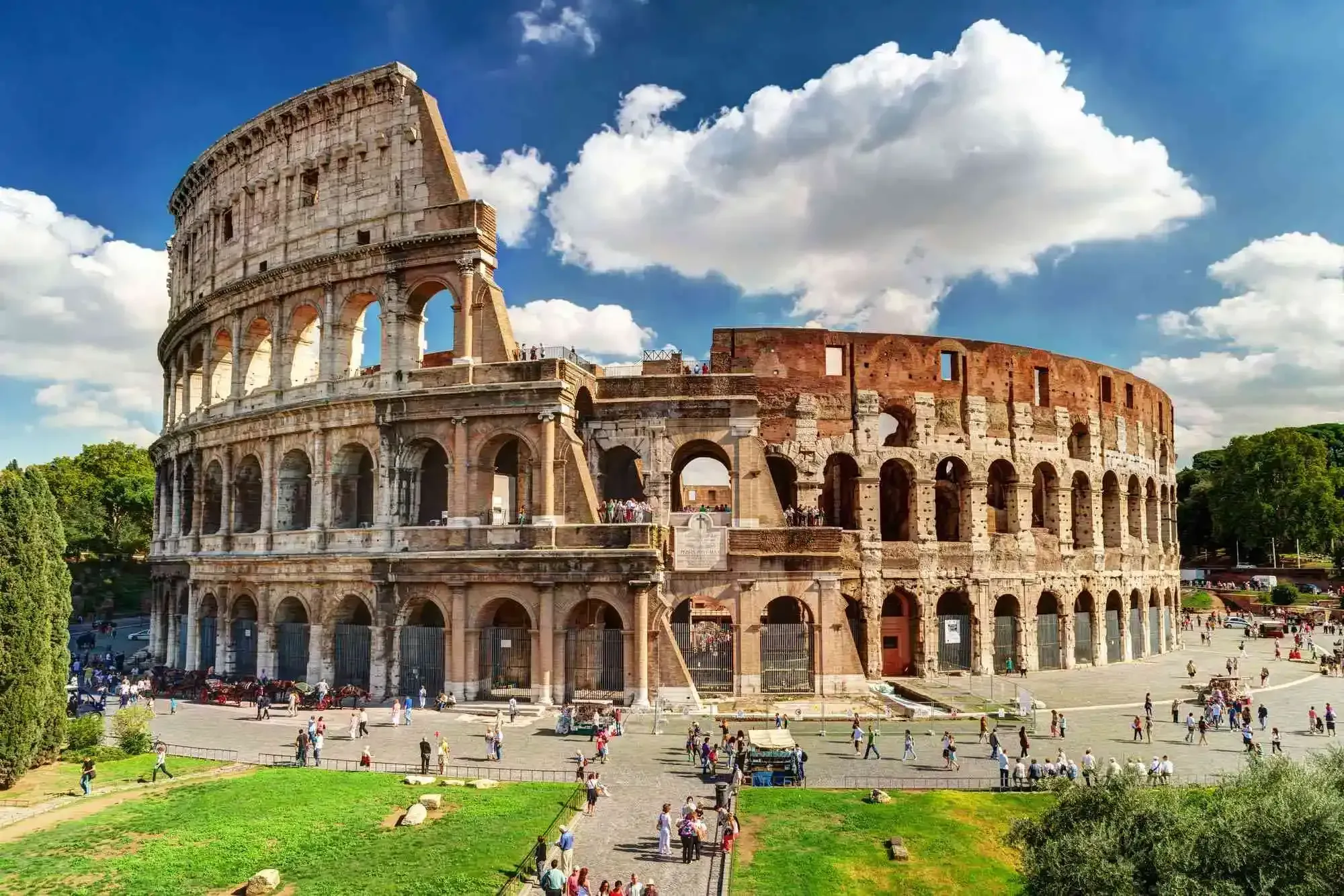 A well-composed, brightly lit image of the Colosseum in Rome, Italy, showcasing its grand architecture against a vivid blue sky with scattered clouds. The foreground includes green grass and numerous tourists, adding scale and life to the scene.