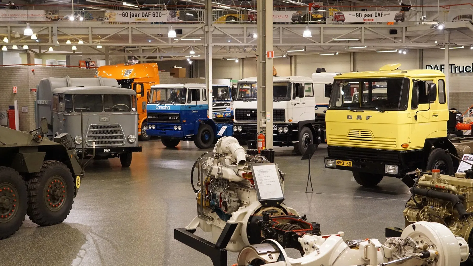 An interior shot of a museum exhibit showcasing several vintage DAF trucks and a large industrial engine on display in the foreground. The hall features exposed industrial ceilings and even lighting.