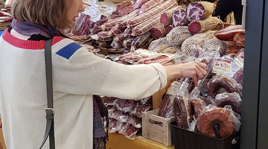An editorial image capturing a person examining or selecting cured meats, such as salami and sausages, displayed at a market stall or deli. The foreground features the person's arm and hand, while the background is filled with a dense array of various packaged and netted meat products.