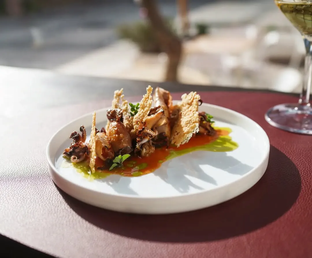 A professionally taken close-up image of a gourmet seafood dish, likely fried octopus or similar small seafood, artfully arranged with crispy elements and vibrant red and green sauces on a white plate. The background is blurred, showing an outdoor or window setting.