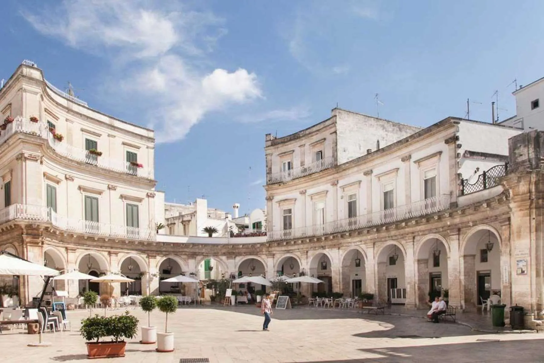 An outdoor, editorial-style photograph of a sunlit historic town square, featuring light-colored multi-story buildings with arched ground levels and decorative balconies. The square is mostly empty, with a few people scattered around, and features outdoor seating areas under the arches. The sky is bright blue with scattered white clouds.