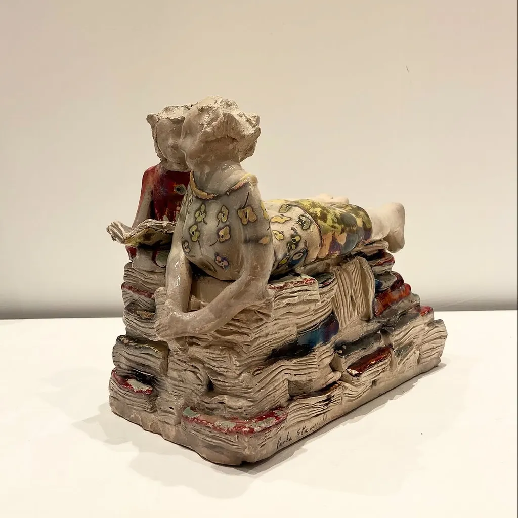 The image captures a ceramic sculpture of figures on books with excellent sharpness and accurate colors. The lighting is soft and even, highlighting the intricate details of the artwork. The primary subject is entirely in focus, contributing to the clarity of the image.