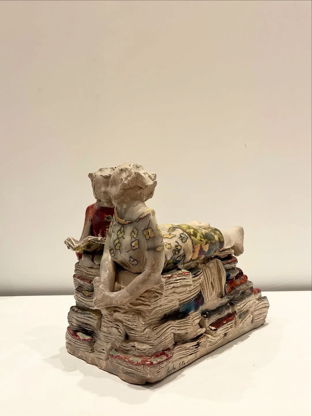 The image captures a ceramic sculpture of figures on books with excellent sharpness and accurate colors. The lighting is soft and even, highlighting the intricate details of the artwork. The primary subject is entirely in focus, contributing to the clarity of the image.