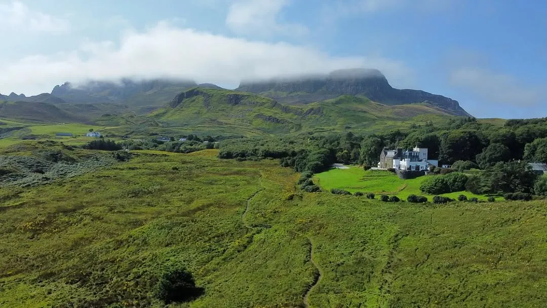 An aerial or high-angle shot showcasing a vast, green, hilly landscape under a partly cloudy sky, with a large white building nestled amongst trees in the midground and imposing mountains in the background. A winding path meanders through the foreground.