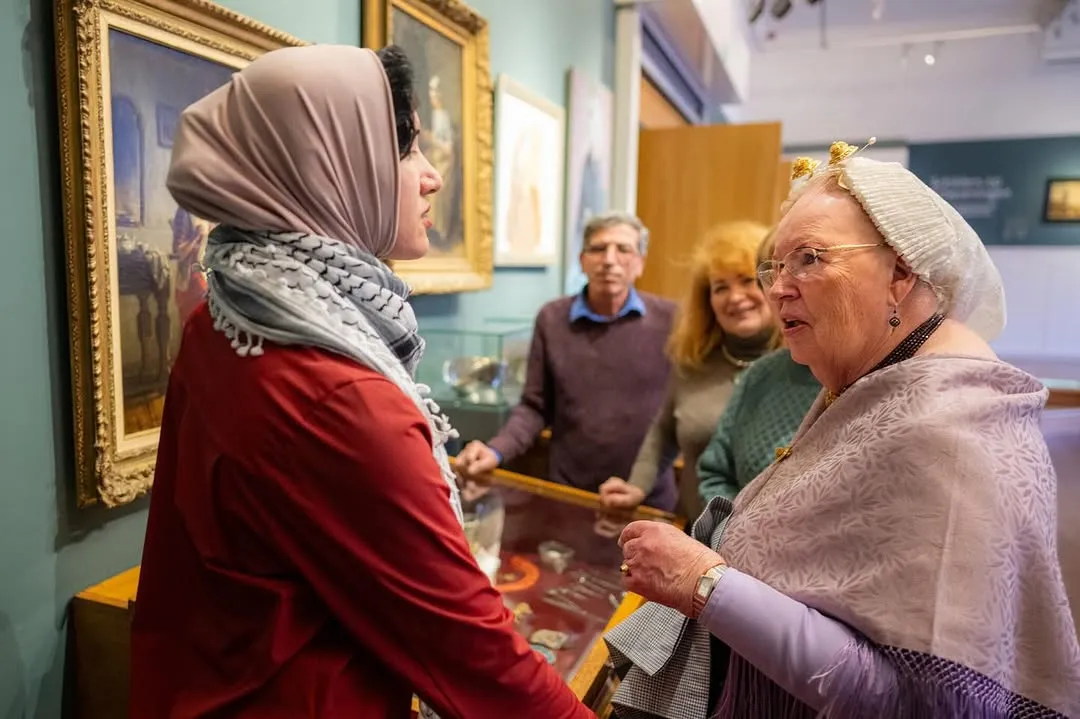 An image capturing an interaction between two women in what appears to be a museum or gallery setting. The foreground subjects are clear and well-focused, engaged in conversation, with other people and exhibits blurred in the background. The scene suggests a cultural or social event.