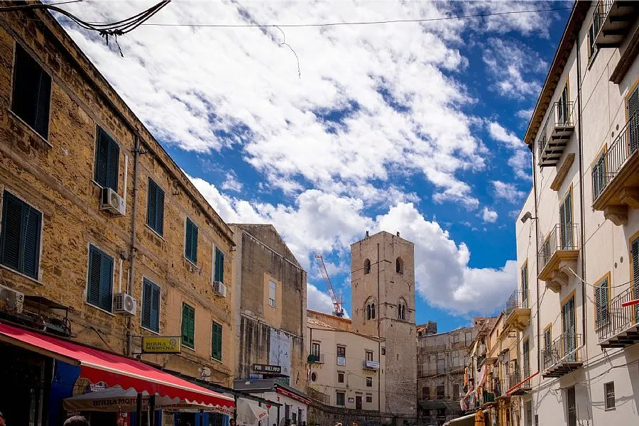 A low-angle view of a narrow street in a historic European city, featuring stone buildings under a bright blue sky with scattered white clouds. A prominent bell tower stands in the background, partially visible.