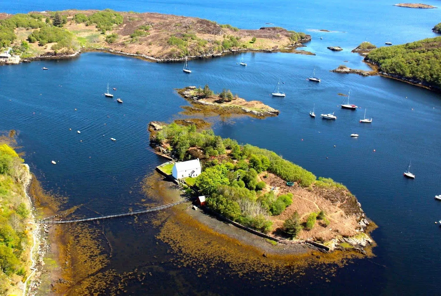 An aerial view of a small, green island featuring a white building, connected by a narrow bridge to a larger landmass. The deep blue water surrounding the islands is dotted with numerous sailing boats and yachts under bright sunlight.