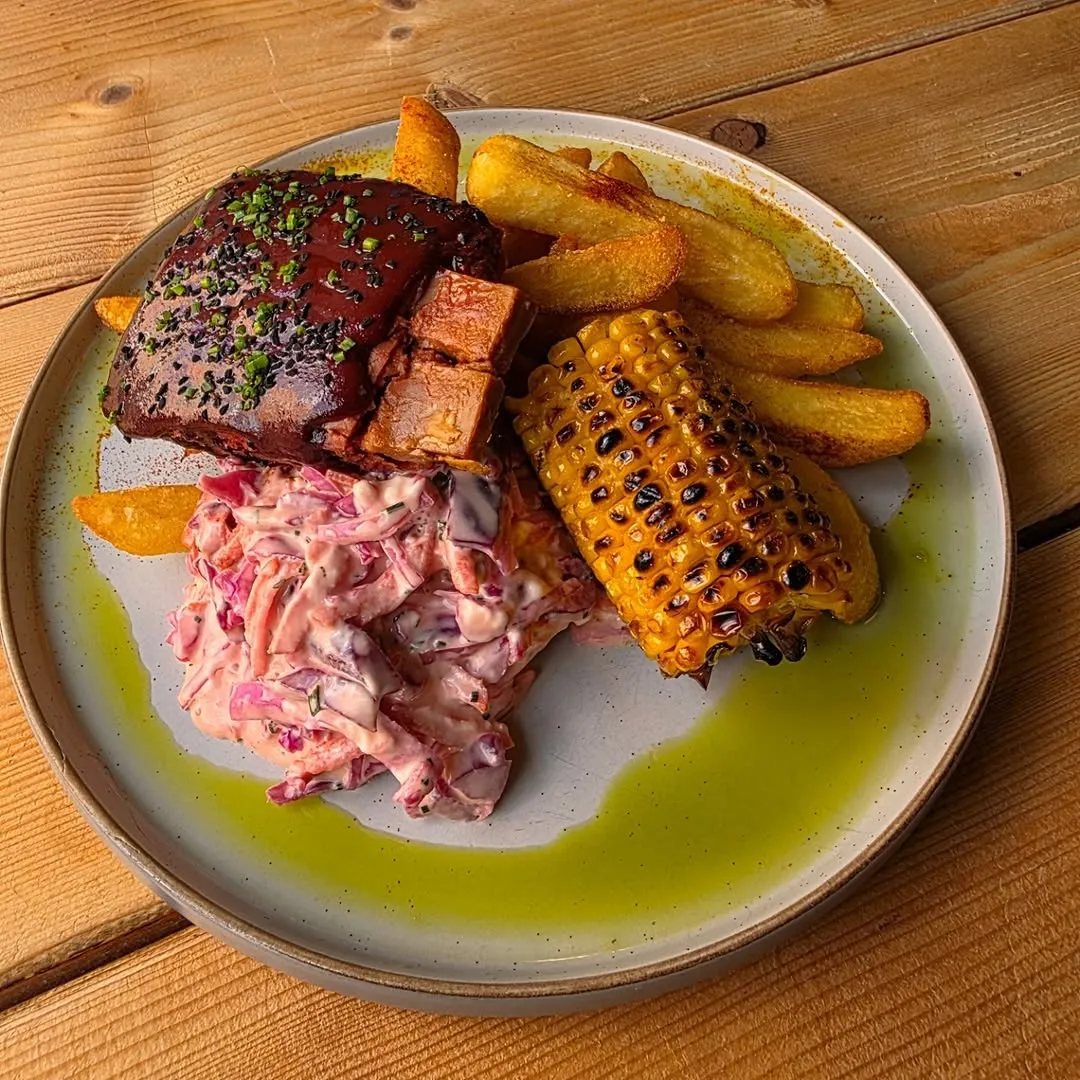 The image is excellently composed, with the dish taking center stage on a simple wooden table, allowing the food to be the sole focus. The lighting is superb, illuminating the textures and details of each component without harshness. All elements, including the ribs, corn, fries, and coleslaw, are exceptionally sharp and in perfect focus, contributing to a high degree of photo-realism. The colors are vibrant and accurate, making the meal look appealing and delicious.