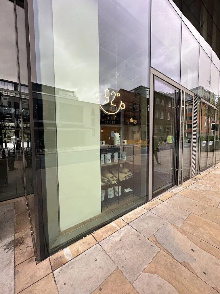 An exterior view of a modern coffee shop storefront with large glass windows reflecting the urban environment and sky. A neon sign reading "92°" is visible inside, above shelves displaying merchandise.