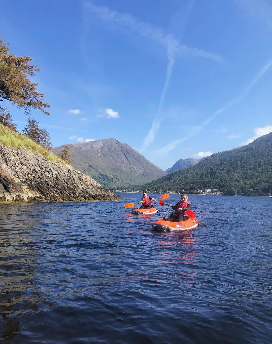 The image captures two individuals kayaking on a calm body of water, possibly a loch or fjord, surrounded by majestic mountains and lush greenery under a clear blue sky. The composition effectively places the subjects within the vast natural landscape.