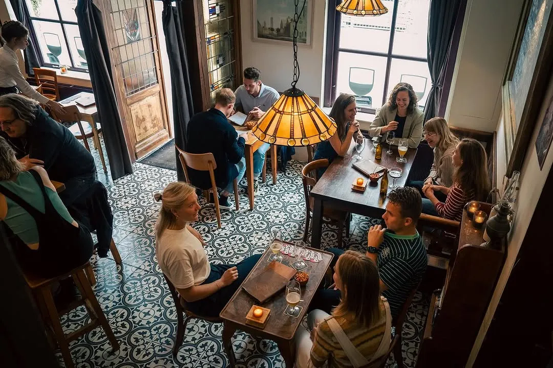 An overhead shot capturing a lively interior scene, possibly a bar or restaurant, with several groups of people socializing at tables. The setting is warm and inviting, characterized by detailed floor tiles and a decorative hanging lamp.