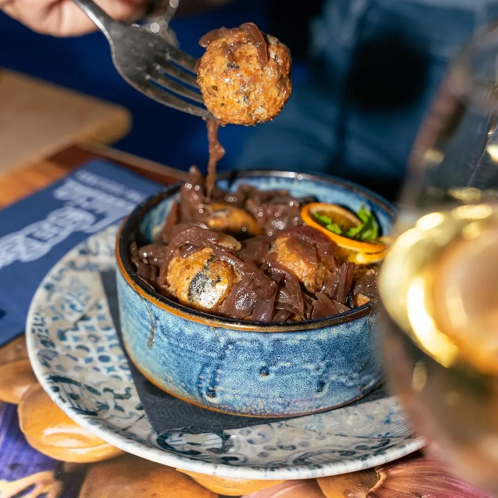 A close-up shot of a fork lifting a meatball covered in a dark sauce with caramelized onions from a blue bowl, with a blurred wine glass in the foreground.