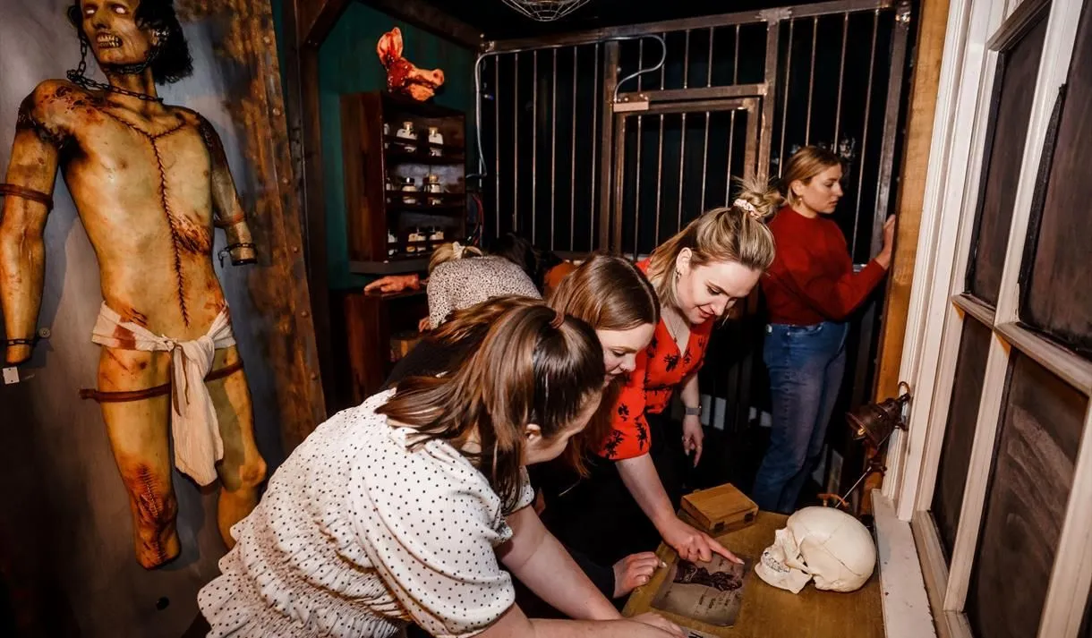 An editorial image showcasing a group of women actively participating in an escape room experience, focused on examining clues on a table, including a book and a skull.