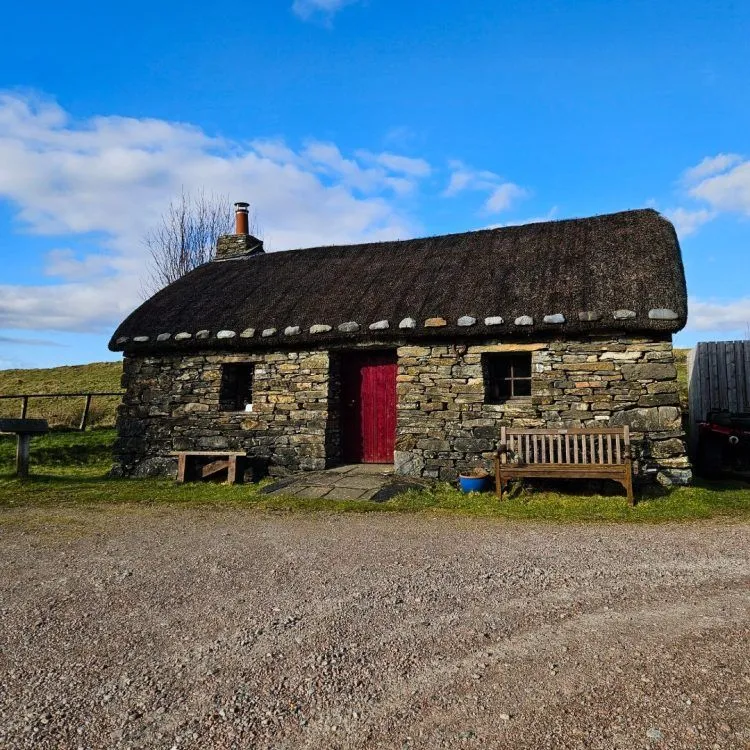 A traditional stone cottage with a thatched roof is centered in the frame under a bright blue sky with wispy clouds. The cottage features a red front door and a small chimney, with wooden benches placed outside. The foreground consists of a gravel area, transitioning to green grass around the cottage.