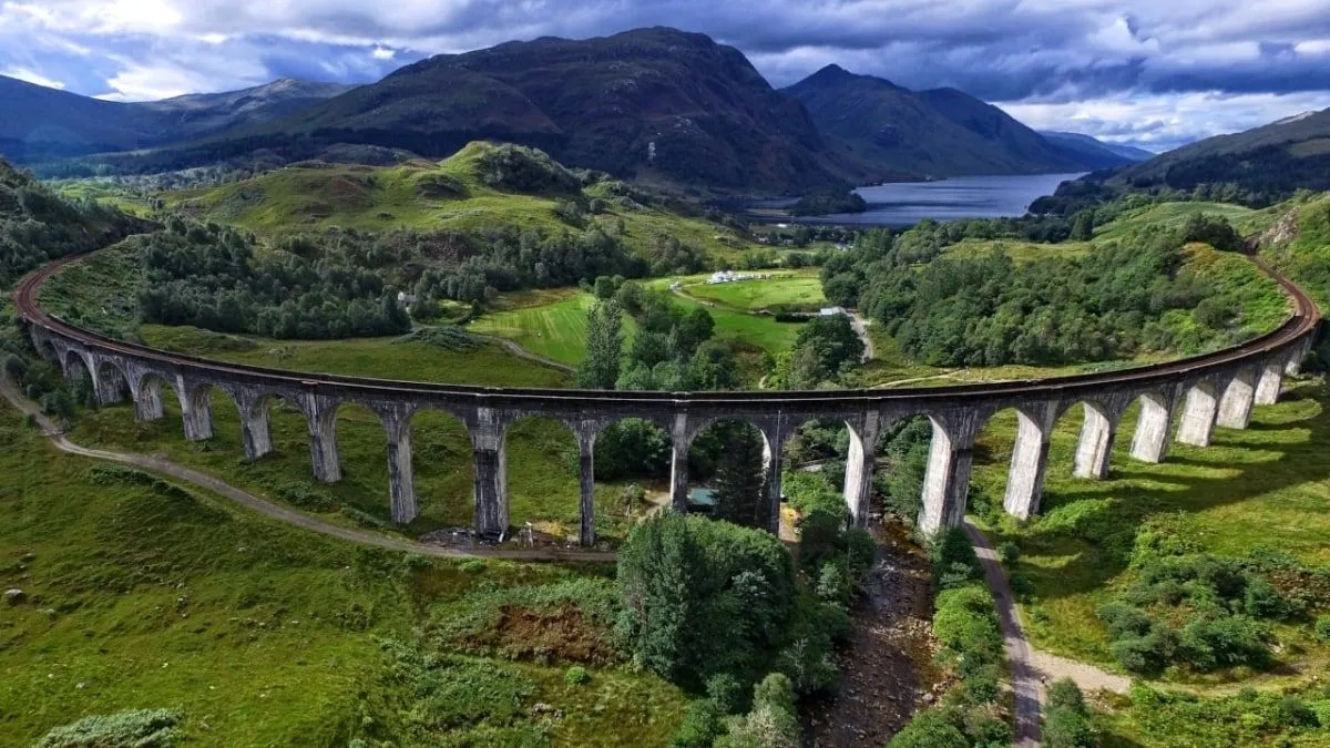 The image captures a scenic landscape featuring the Glenfinnan Viaduct amidst rolling green hills, distant mountains, and a body of water.