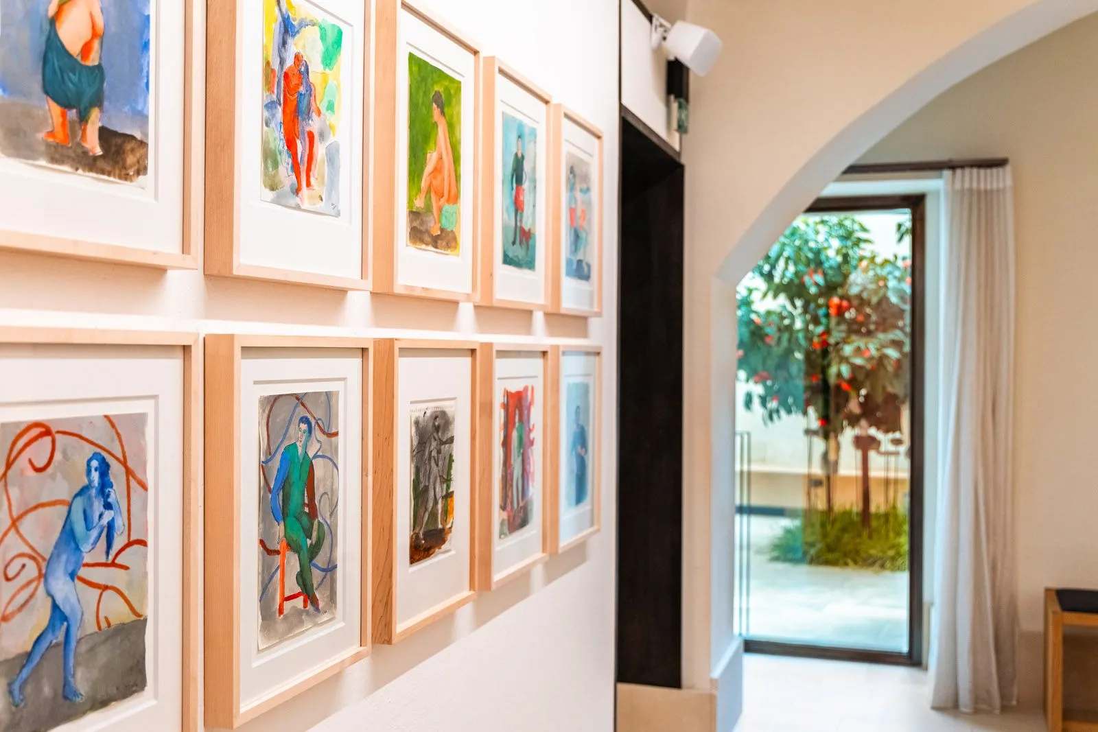 A well-lit, editorial-style photograph of a hallway or art gallery. The left side of the frame showcases a white wall adorned with two rows of framed, colorful abstract artworks. The right side features an arched doorway leading to an outdoor courtyard with lush greenery and a tree, creating a sense of depth. The image employs a shallow depth of field, keeping the artworks in sharp focus while the background is softly blurred.