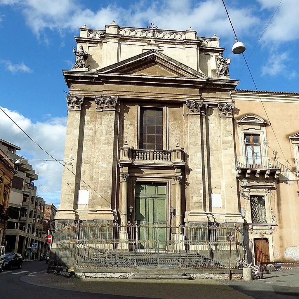 A sunlit exterior view of an ornate, classical-style stone building, possibly a church, featuring a large green main door, an elevated balcony, and intricate architectural details. The building occupies the central frame, with a metal fence and street in the foreground, and other urban structures visible on the sides. A blue sky with some clouds is overhead, though power lines and a hanging street lamp cross the upper portion of the frame.