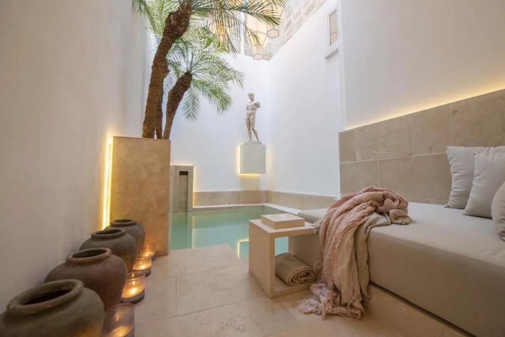 A serene and aesthetically pleasing image of an indoor spa or relaxation area, featuring a small pool, palm trees, a classical statue, and comfortable seating with soft lighting.