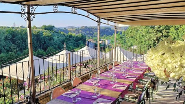 An inviting outdoor dining setup on a terrace, overlooking a lush, green landscape under clear skies. A long table is meticulously set with vibrant purple and gold runners, empty glasses, and napkins.