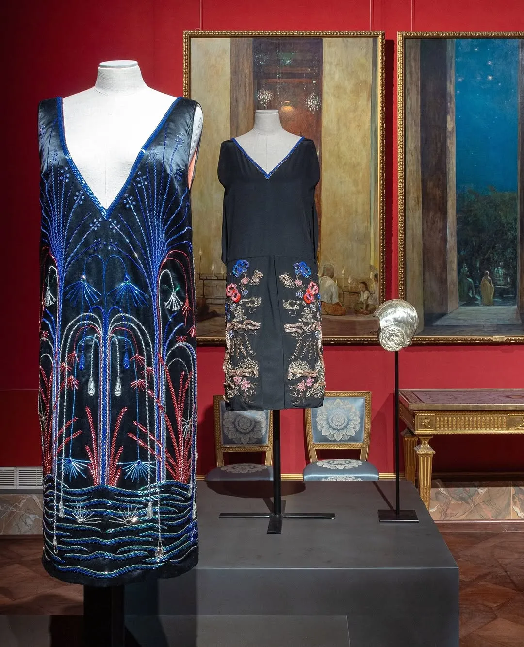 Two intricately decorated dresses displayed on mannequins in what appears to be a museum or exhibition setting, with a large painting in the background and ornate furniture.