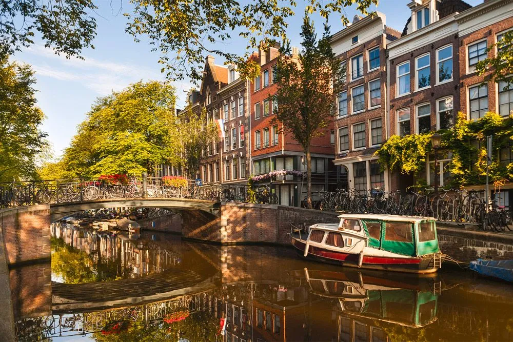 A picturesque scene of an Amsterdam canal, featuring traditional Dutch canal houses, a bridge laden with bicycles, and several canal boats, all beautifully reflected in the water under warm, golden hour lighting.