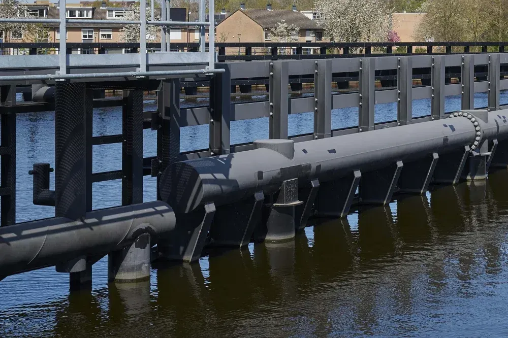 An image capturing a modern water control structure or sluice gate on a canal. The dark metal gates and machinery are prominently featured, with reflections visible in the calm water below. In the background, there are houses and trees, some of which appear to be blooming.