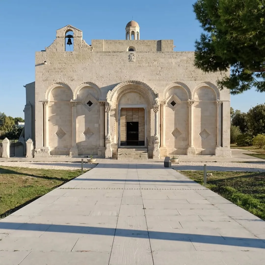 The image depicts a grand stone building with classical architectural elements, including arched facades and columns, centered under a bright blue sky. A wide, light-colored paved pathway extends from the foreground directly to the building's entrance, flanked by green grass and distant trees.