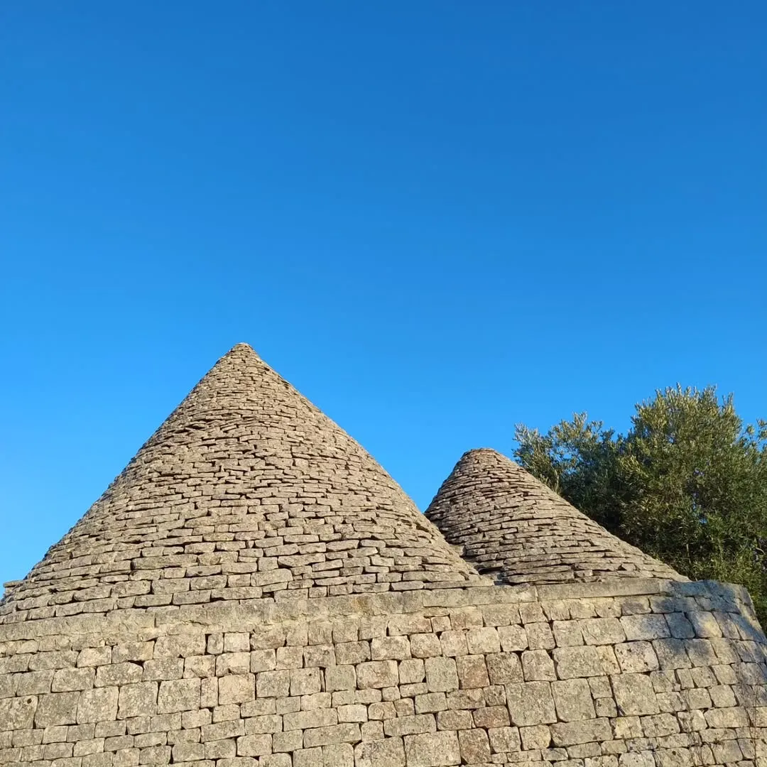 The image prominently features two traditional Trulli houses with their distinctive conical stone roofs against a clear blue sky. The stone work is sharply detailed and well-lit, contributing to a strong sense of realism. The composition effectively highlights the unique architectural elements.