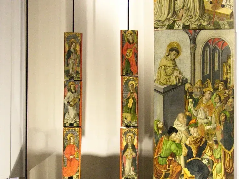 A photograph showcasing several historical panel paintings, likely religious art, displayed in a museum or gallery setting. The image features two vertical columns of smaller, individual painted panels on the left, and a larger, multi-figure panel painting on the right, which is partially cropped.