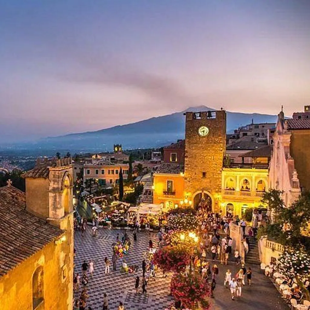 An elevated view of a lively town square and surrounding buildings at twilight. The scene features a prominent clock tower, a church roof in the foreground, and a bustling square filled with people. In the background, a coastline extends towards mountains under a colorful sky.