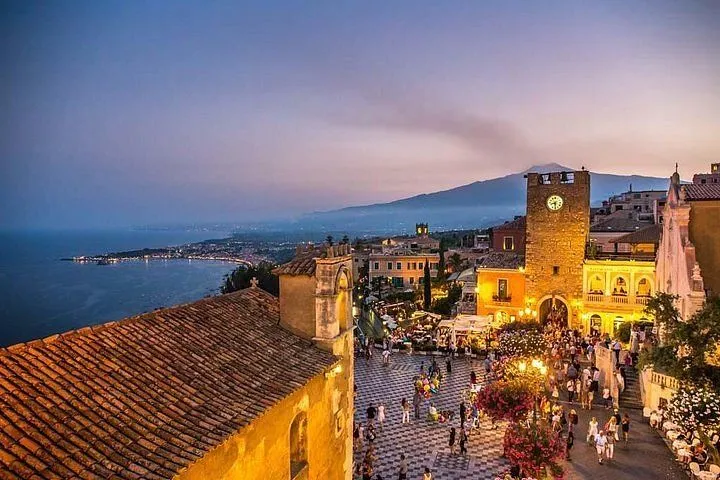 An elevated view of a lively town square and surrounding buildings at twilight. The scene features a prominent clock tower, a church roof in the foreground, and a bustling square filled with people. In the background, a coastline extends towards mountains under a colorful sky.