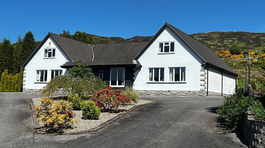 A front view of a white stucco house with a dark roof and dark framed windows, surrounded by green and red bushes, with a paved driveway in the foreground and mountains under a clear blue sky in the background.