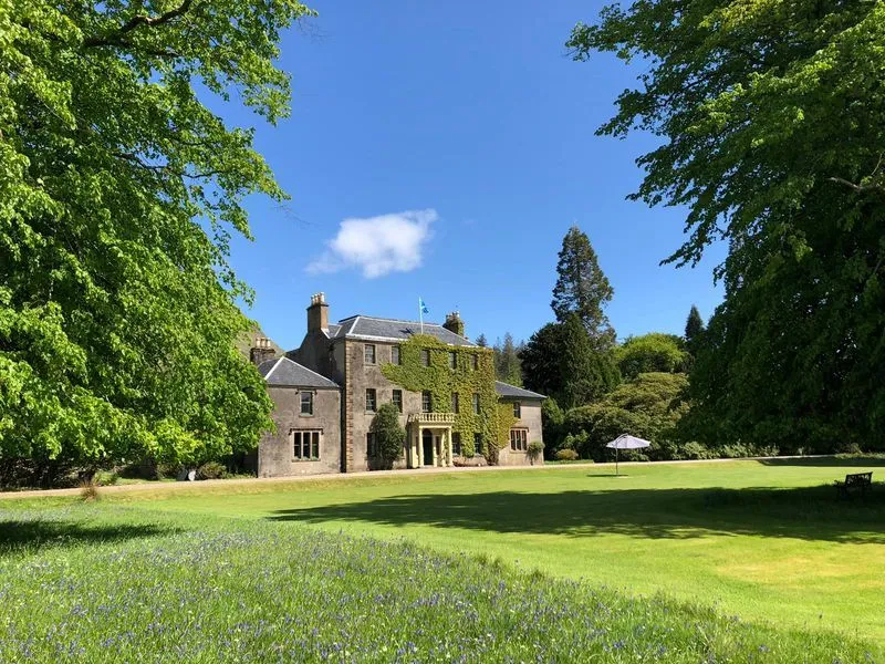 A grand, ivy-covered historic building stands majestically in a lush green landscape under a bright blue sky. The scene is framed by vibrant green trees, and the foreground features a sprawling lawn with subtle blue wildflowers.