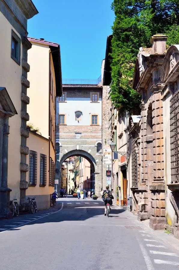 A sunlit street scene in what appears to be a historic European town, featuring old stone buildings, an archway, and people including one on a bicycle. The composition effectively leads the eye down the street.