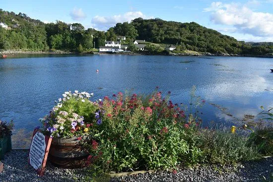 This image showcases a beautiful coastal landscape with vibrant flowers in the foreground, leading to calm blue waters and white houses nestled against a forested hill in the distance. The composition uses depth effectively, drawing the viewer's eye through the scene.
