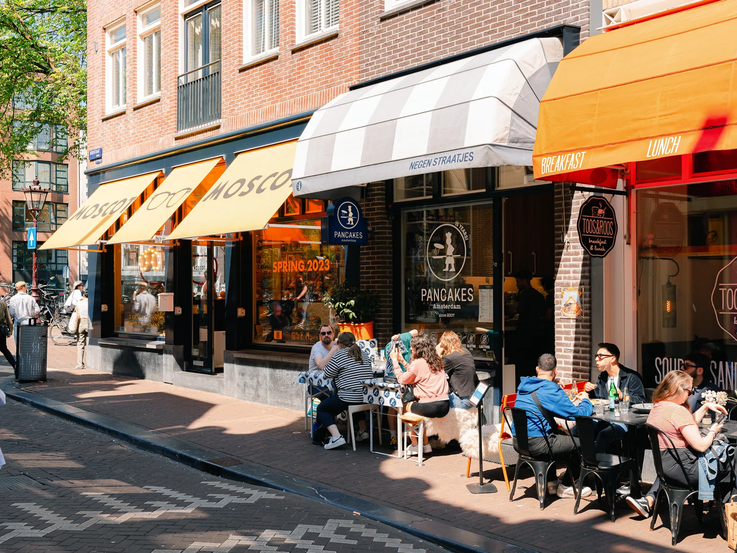 An inviting street scene featuring outdoor dining areas in front of several businesses, including a pancake restaurant. People are seated at tables under awnings, enjoying the sunny weather. The architecture is typical of a European city street, with brick buildings and distinct storefronts.