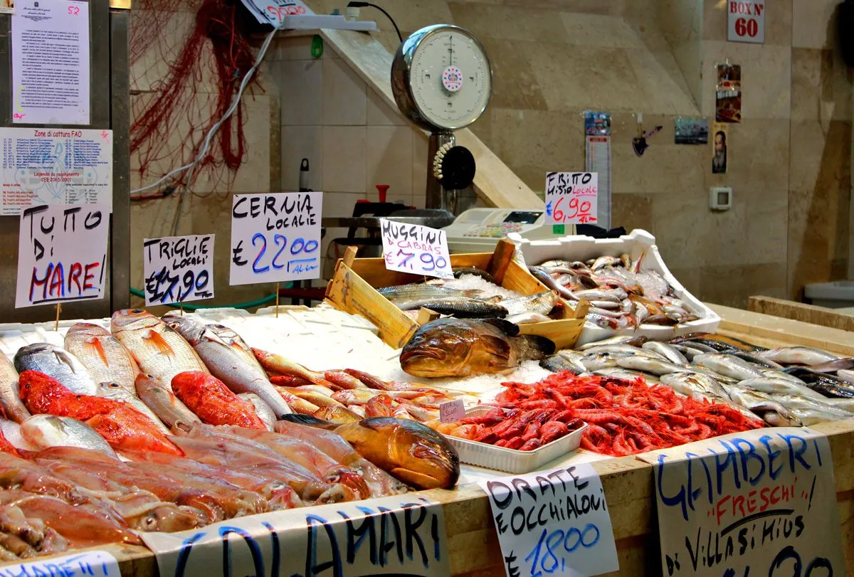 An editorial-style image showcasing a vibrant fish market stall filled with various types of fresh fish and seafood on ice, with pricing signs in Italian. A large vintage-style scale is visible in the background.