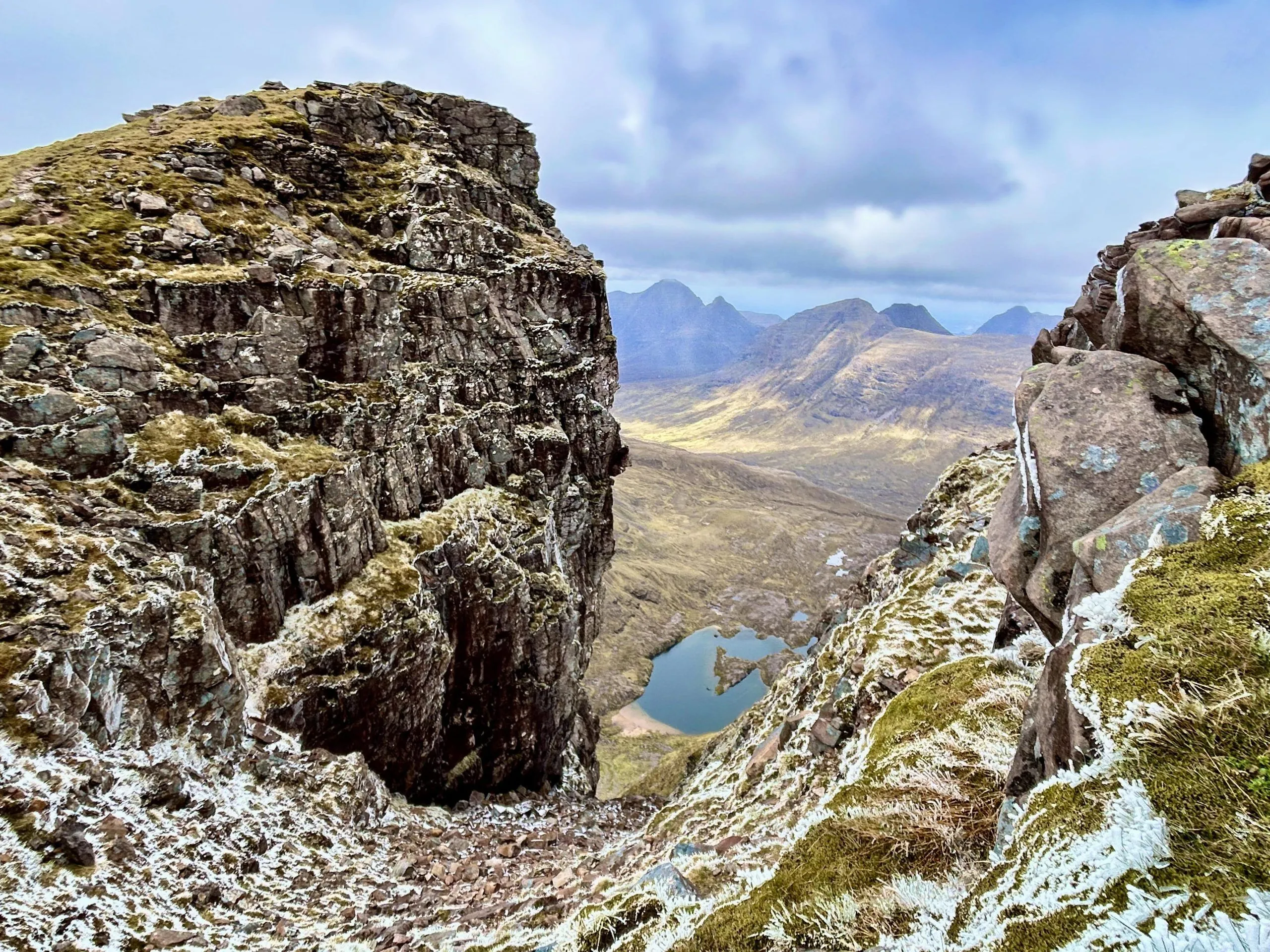 An editorial quality image showcasing a dramatic mountain landscape with towering cliffs in the foreground that frame a deep valley, a small lake, and distant mountains under a cloudy sky. Patches of snow and frost are visible on the rugged terrain.